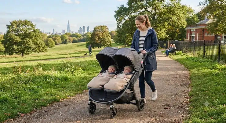 A side-by-side double pram suitable for newborn twins, walking along a British park path.